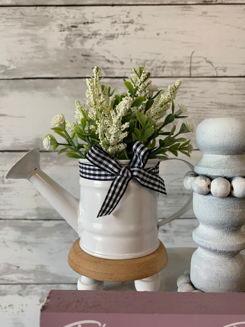 May include: A white metal watering can filled with white and green faux flowers, accented with a black and white checkered bow. The watering can sits on a small wooden stand with white legs. Decorative home decor.