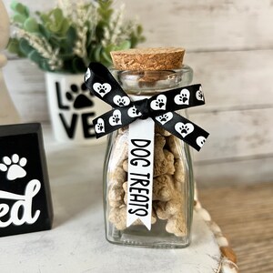 May include: A clear glass jar filled with dog treats, tied with a black ribbon with white paw prints. The ribbon has a white tag that says "DOG TREATS".