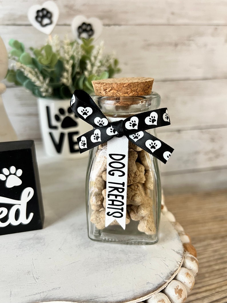 May include: A clear glass jar filled with dog treats, tied with a black ribbon with white paw prints. The ribbon has a white tag that says "DOG TREATS".