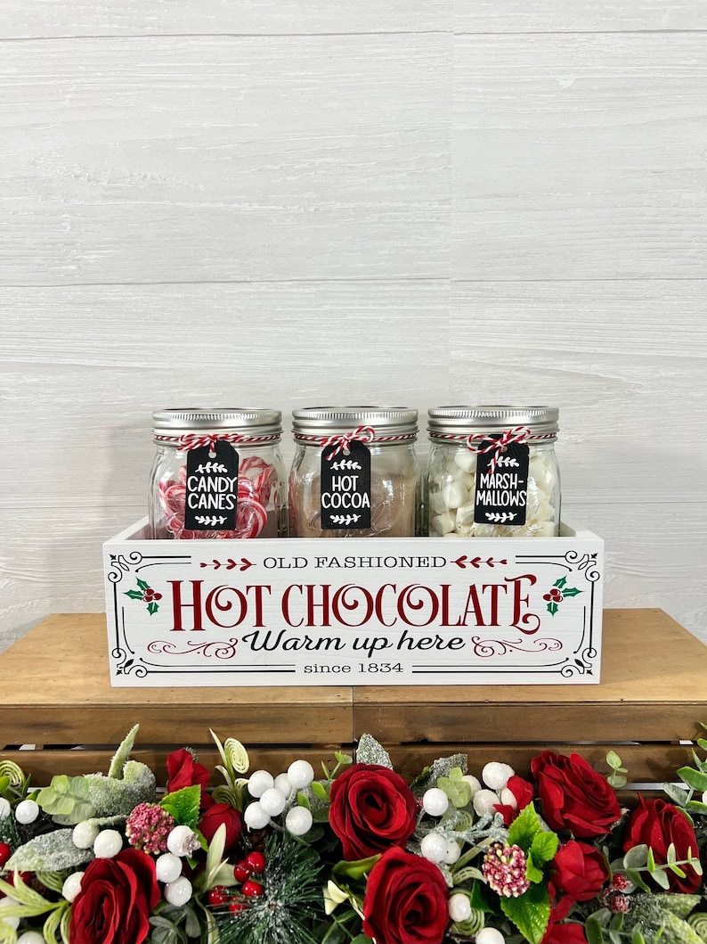 May include: A white wooden box with the text "Old Fashioned Hot Chocolate Warm up here since 1834" on the front. The box contains three glass jars filled with candy canes, hot cocoa mix, and marshmallows.
