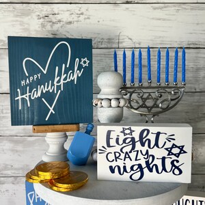 May include: A white tiered tray decorated for Hanukkah with blue and white decorations. The top tier has a blue sign that says "Happy Hanukkah" with a white heart and star of David. The middle tier has a silver menorah with blue candles, a blue dreidel, and a white sign that says "Eight Crazy Nights" with a star of David. The bottom tier has a blue sign with a star of David and a pile of gold chocolate coins.