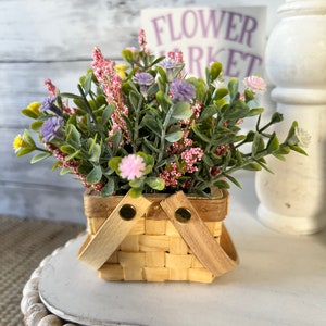 May include: A small wicker basket with wooden handles filled with a bouquet of pink, purple, and yellow flowers. The basket is sitting on a white shelf with a sign that reads "Flower Market".