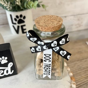 May include: A glass jar filled with dog treats, tied with a black and white ribbon with paw print and heart designs. The jar has a cork lid and a white label that reads "DOG TREATS".