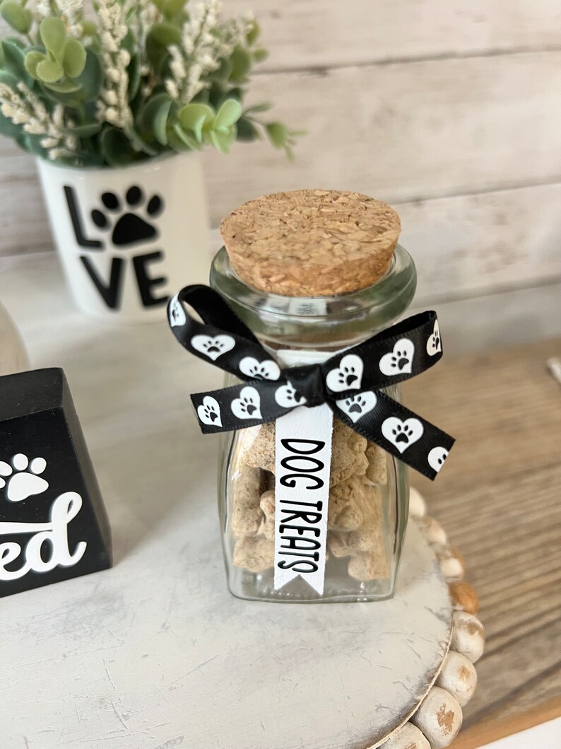 May include: A glass jar filled with dog treats, tied with a black and white ribbon with paw print and heart designs. The jar has a cork lid and a white label that reads "DOG TREATS".