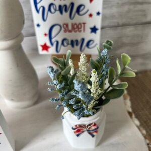 Pode incluir: Um vaso de cer&acirc;mica branco com um la&ccedil;o vermelho, branco e azul est&aacute; sobre uma superf&iacute;cie branca. O vaso cont&eacute;m um buqu&ecirc; de flores artificiais azuis e verdes. Um letreiro de madeira com o texto "my home sweet home" em letras azuis e vermelhas &eacute; vis&iacute;vel no fundo.