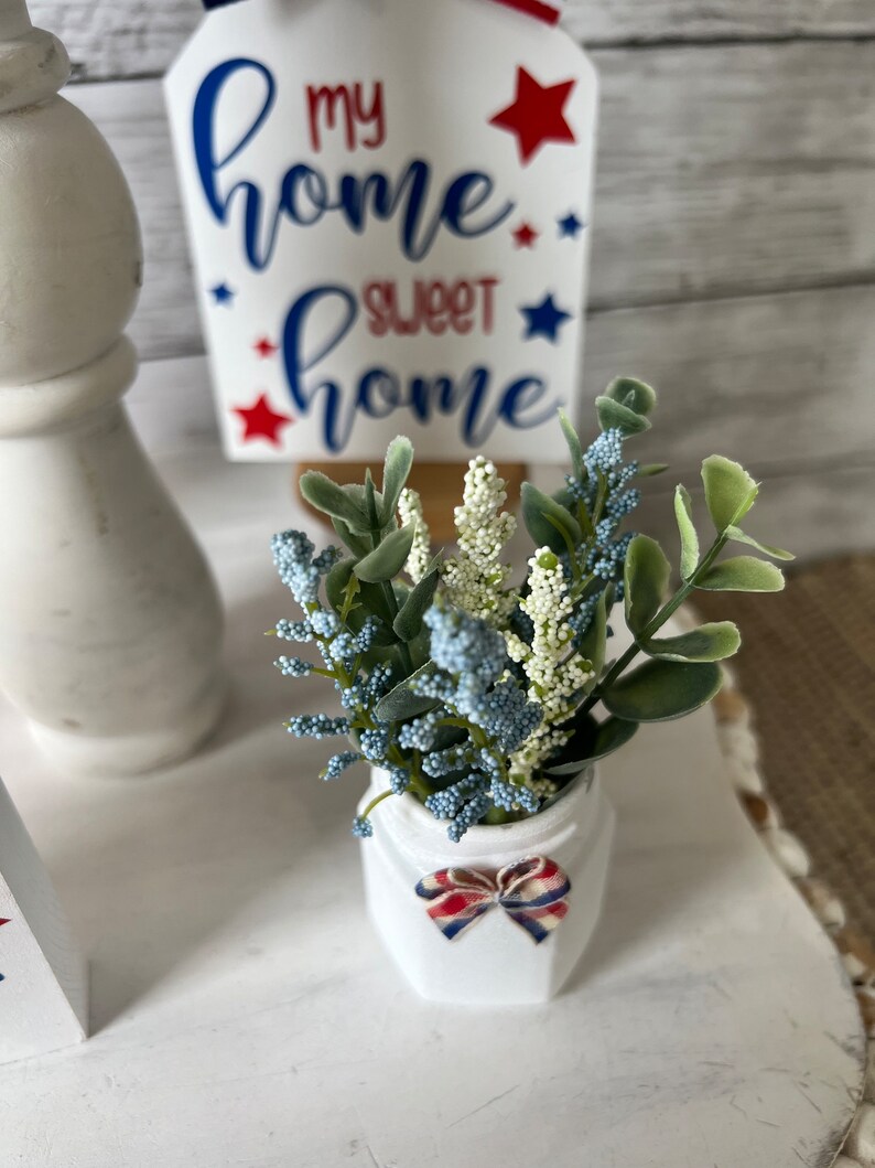 Pode incluir: Um vaso de cer&acirc;mica branco com um la&ccedil;o vermelho, branco e azul est&aacute; sobre uma superf&iacute;cie branca. O vaso cont&eacute;m um buqu&ecirc; de flores artificiais azuis e verdes. Um letreiro de madeira com o texto "my home sweet home" em letras azuis e vermelhas &eacute; vis&iacute;vel no fundo.