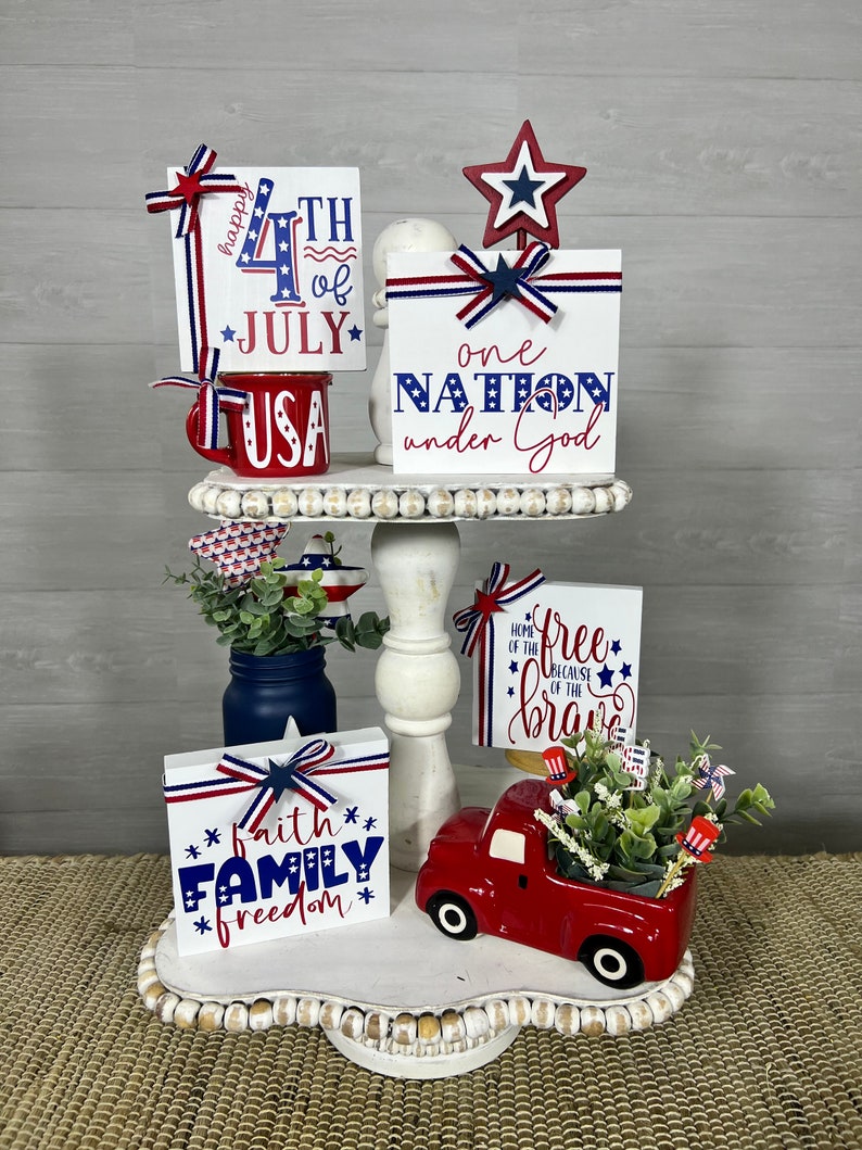 May include: A tiered tray decorated for the 4th of July with red, white, and blue wooden signs. The signs say "Happy 4th of July", "USA", "One Nation Under God", "Home of the Free Because of the Brave", and "Faith Family Freedom". There is a red truck planter with greenery and small American flags.