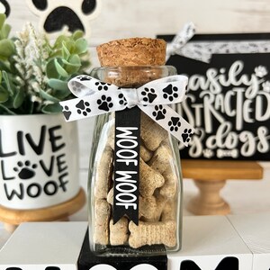 May include: A glass jar filled with dog treats, tied with a white ribbon with black paw prints. The jar has a cork lid and a black tag that says "Woof Woof". The jar is sitting on a white block with the word "Home" in black letters.