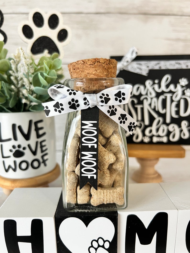 May include: A glass jar filled with dog treats, tied with a white ribbon with black paw prints. The jar has a cork lid and a black tag that says "Woof Woof". The jar is sitting on a white block with the word "Home" in black letters.