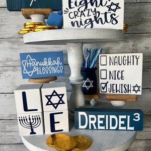 May include: A tiered tray decorated for Hanukkah with blue and white decorations. The top tier features a sign that says "Eight Crazy Nights" and a menorah with blue candles. The middle tier has a sign that says "Hanukkah Blessings", a dreidel, and a sign that says "Naughty, Nice, Jewish". The bottom tier has a sign that says "Happy Hanukkah" and a dreidel.