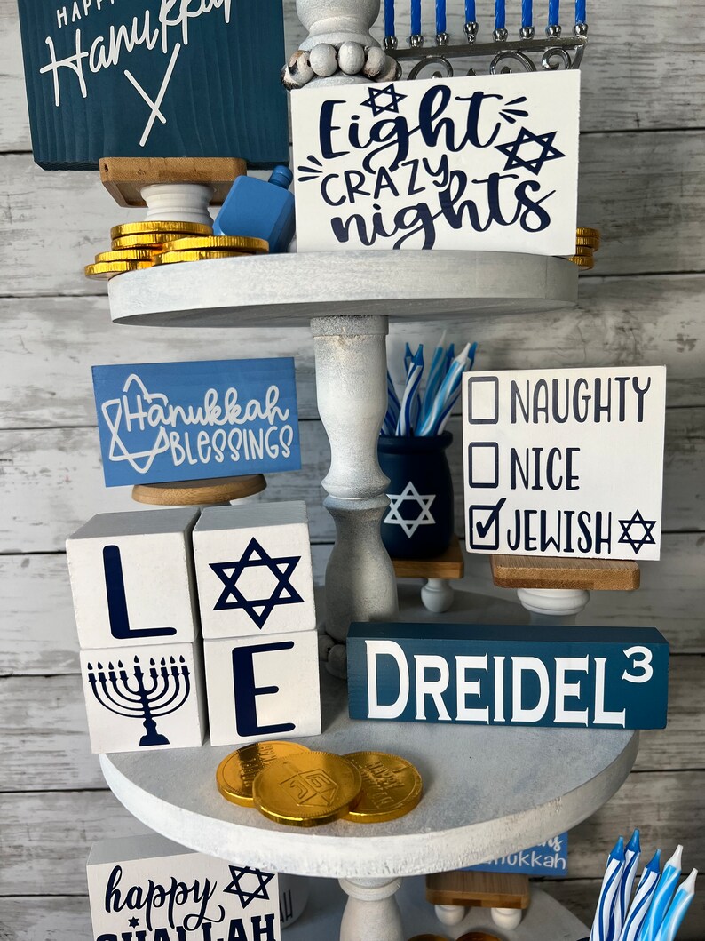 May include: A tiered tray decorated for Hanukkah with blue and white decorations. The top tier features a sign that says "Eight Crazy Nights" and a menorah with blue candles. The middle tier has a sign that says "Hanukkah Blessings", a dreidel, and a sign that says "Naughty, Nice, Jewish". The bottom tier has a sign that says "Happy Hanukkah" and a dreidel.