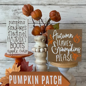 May include: Three wooden signs with fall-themed text. The top sign says "pumpkins bonfires flannel hayrides boots apple cider". The middle sign says "Autumn leaves pumpkins please". The bottom sign says "Pumpkin Patch corn maze hayrides cider".