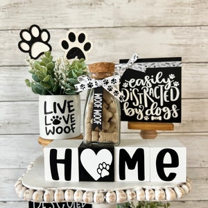 May include: A tiered tray decorated with dog-themed home decor. The top tier features a glass jar filled with dog treats, a white ceramic mug with the words "Live Love Woof", and two black and white paw print decorations. The middle tier features four white wooden blocks spelling "HOME" with a black paw print heart in the center. The bottom tier features a black and white sign with the text "Rescued is my favorite breed" and a small potted plant.