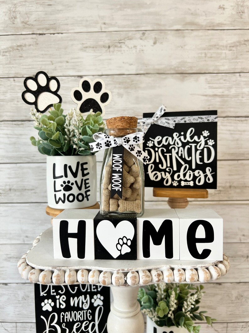 May include: A tiered tray decorated with dog-themed home decor. The top tier features a glass jar filled with dog treats, a white ceramic mug with the words "Live Love Woof", and two black and white paw print decorations. The middle tier features four white wooden blocks spelling "HOME" with a black paw print heart in the center. The bottom tier features a black and white sign with the text "Rescued is my favorite breed" and a small potted plant.