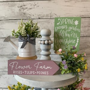 May include: A display of spring-themed decorative items. Includes a white watering can with a black and white checkered bow, a wooden sign that reads "Flower Farm", and a green sign with the word "Spring". Artificial flowers are also present.