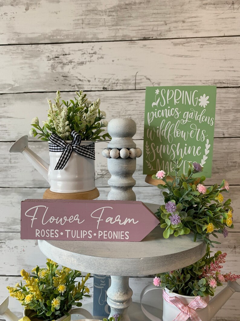May include: A display of spring-themed decorative items. Includes a white watering can with a black and white checkered bow, a wooden sign that reads "Flower Farm", and a green sign with the word "Spring". Artificial flowers are also present.