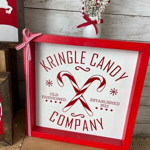 May include: Red wooden sign with white text that reads "Kringle Candy Company" with a red and white candy cane design. The text also reads "Old Fashioned" and "Established 1822".