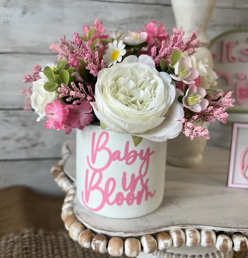 May include: A white ceramic planter with pink lettering that reads "Baby in Bloom" is filled with a bouquet of pink and white flowers.