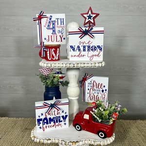 May include: A tiered tray decorated for the 4th of July with red, white, and blue wooden signs. The signs say "happy 4th of JULY", "one NATION under God", "faith FAMILY freedom", and "HOME OF THE free BECAUSE OF THE brave".