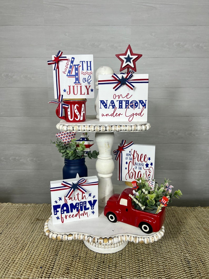 May include: A tiered tray decorated for the 4th of July with red, white, and blue wooden signs. The signs say "happy 4th of JULY", "one NATION under God", "faith FAMILY freedom", and "HOME OF THE free BECAUSE OF THE brave".