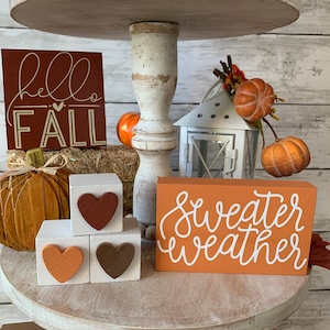 May include: A three-tiered wooden stand decorated with fall-themed items. The top tier features a sign that says "hello fall" and a small pumpkin. The middle tier has a sign that says "sweater weather" and three wooden blocks with hearts. The bottom tier has a sign that says "farm fresh" and a small pumpkin.