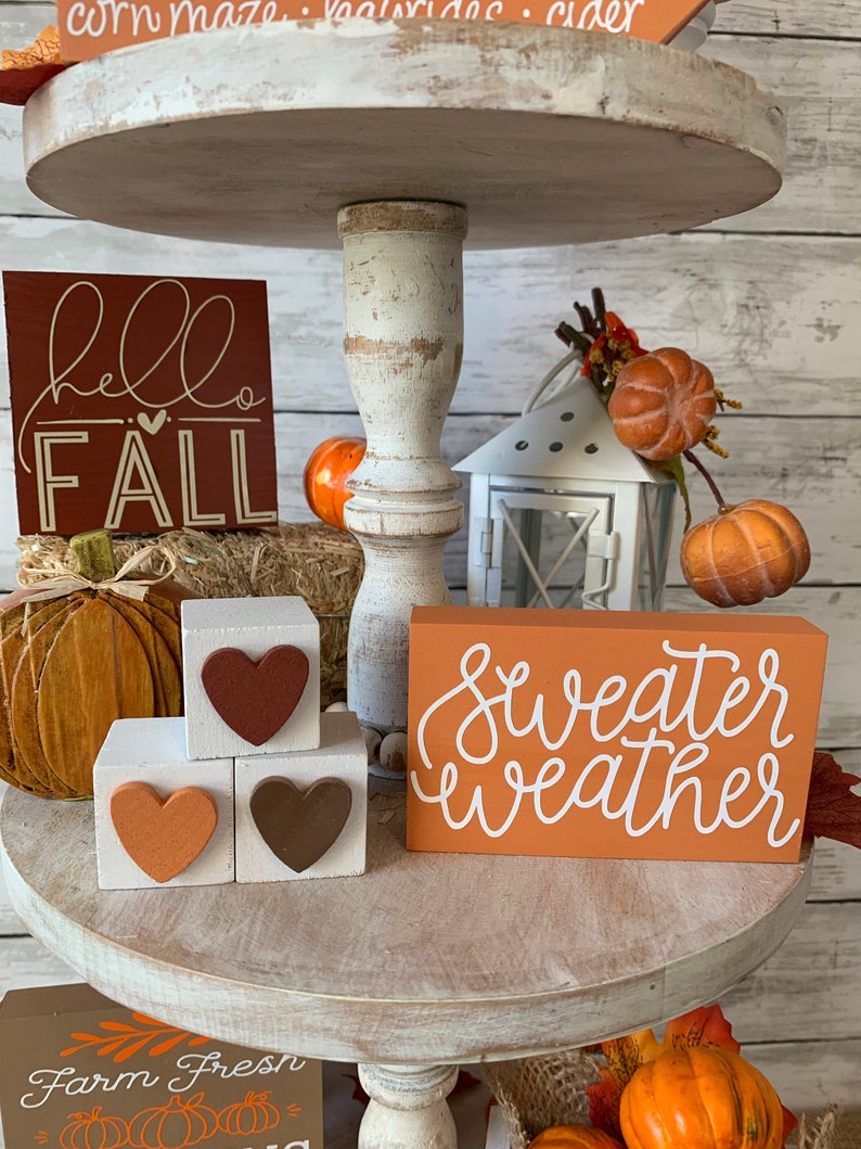 May include: A three-tiered wooden stand decorated with fall-themed items. The top tier features a sign that says "hello fall" and a small pumpkin. The middle tier has a sign that says "sweater weather" and three wooden blocks with hearts. The bottom tier has a sign that says "farm fresh" and a small pumpkin.