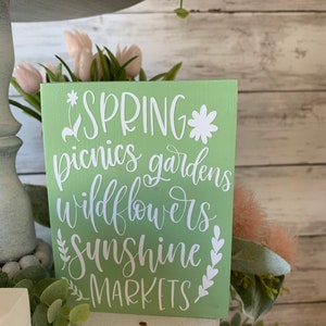 Pu&ograve; includere: Un cartello di legno verde chiaro con lettere bianche che recita "SPRING picnics gardens wildflowers sunshine MARKETS".