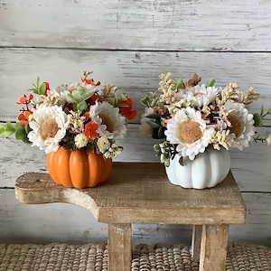 May include: Two floral arrangements in pumpkin-shaped vases. One is orange, the other white. Each arrangement features white and tan flowers with green and orange accents, ideal for autumn decoration.