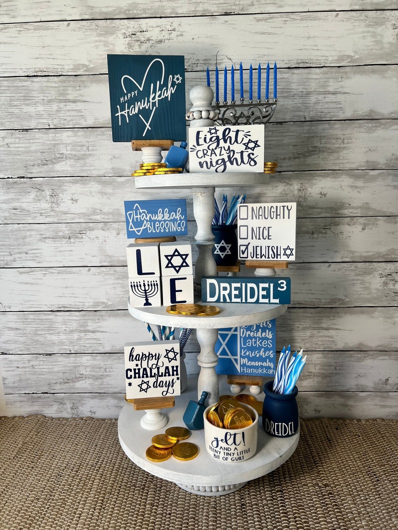 May include: A tiered tray decorated for Hanukkah with blue and white signs, a dreidel, and gold chocolate coins. The signs say "Happy Hanukkah", "Eight Crazy Nights", "Hanukkah Blessings", "L'E", "Happy Challah Days", "Dreidels, Latkes, Knishes, Menorahs, Hanukkah", "Naughty, Nice, Jewish", "Dreidel 3", and "Gelt! And a Tiny Little Bit of Guilt".