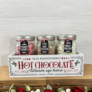 May include: A white wooden box with the words "Old Fashioned Hot Chocolate Warm up here since 1834" printed on the front. Three glass jars filled with candy canes, hot cocoa mix, and marshmallows are sitting inside the box.