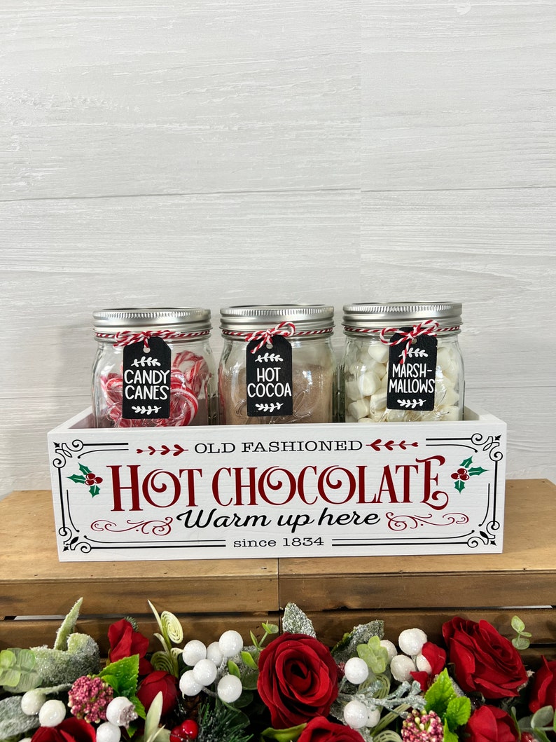 May include: A white wooden box with the words "Old Fashioned Hot Chocolate Warm up here since 1834" printed on the front. Three glass jars filled with candy canes, hot cocoa mix, and marshmallows are sitting inside the box.