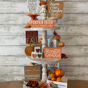 May include: A tiered tray decorated for fall with various signs and pumpkins. The signs include "Pumpkins Bonfires Flannel Hayrides Boots Apple Cider", "Autumn Leaves Pumpkins Please", "Pumpkin Patch Corn Maze Hayrides Cider", "Hello Fall", "Sweater Weather", "Spice Everything", "Farm Fresh Pumpkins Picked Daily", "Jump in the Leaves", and "Pumpkins 25c".
