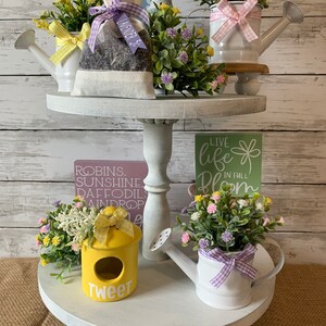May include: A three-tiered, white, distressed decorative tray with spring-themed decor. Includes miniature watering cans with flowers, a yellow birdhouse with "Tweet" text, and a lavender sachet. The background is a white wood-paneled wall.