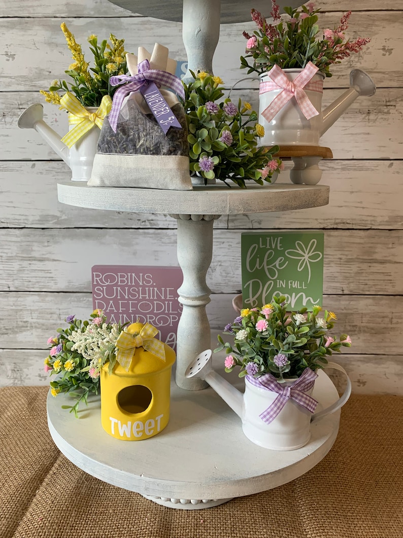 May include: A three-tiered, white, distressed decorative tray with spring-themed decor. Includes miniature watering cans with flowers, a yellow birdhouse with "Tweet" text, and a lavender sachet. The background is a white wood-paneled wall.