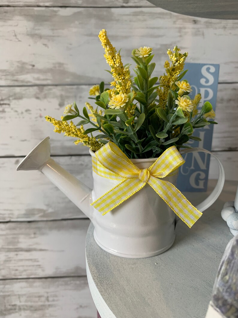 May include: A small white metal watering can filled with yellow and green faux flowers. A yellow and white checkered ribbon is tied around the watering can. A blue sign with the word "SPRING" is in the background.