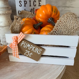 May include: A white wooden crate filled with orange pumpkins and burlap. A brown sign with white text reads "PUMPKINS 25&cent;".