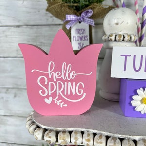 K&ouml;nnte beinhalten: Ein rosa, tulpenf&ouml;rmiges Schild aus Holz mit dem Text "hello spring" in wei&szlig;en Buchstaben. Das Schild steht auf einem mehrstufigen Regal mit wei&szlig;en Perlen am Rand. Es gibt einen kleinen Blumenstrau&szlig; in einer mit Sackleinen umwickelten Vase und einen wei&szlig;en Keramiksockel mit einem lila-wei&szlig; gestreiften Strohhalm.