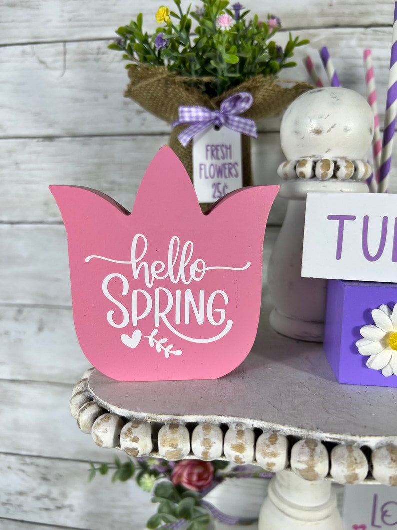 K&ouml;nnte beinhalten: Ein rosa, tulpenf&ouml;rmiges Schild aus Holz mit dem Text "hello spring" in wei&szlig;en Buchstaben. Das Schild steht auf einem mehrstufigen Regal mit wei&szlig;en Perlen am Rand. Es gibt einen kleinen Blumenstrau&szlig; in einer mit Sackleinen umwickelten Vase und einen wei&szlig;en Keramiksockel mit einem lila-wei&szlig; gestreiften Strohhalm.