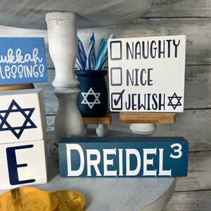 May include: A tiered tray decorated for Hanukkah with blue and white decorations. The tray features a dreidel, a menorah, and a sign that says "Naughty, Nice, Jewish".