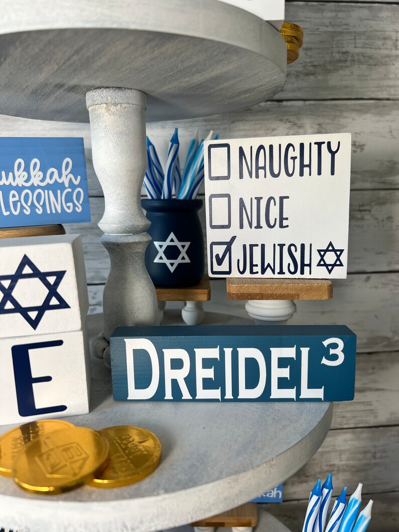 May include: A tiered tray decorated for Hanukkah with blue and white decorations. The tray features a dreidel, a menorah, and a sign that says "Naughty, Nice, Jewish".