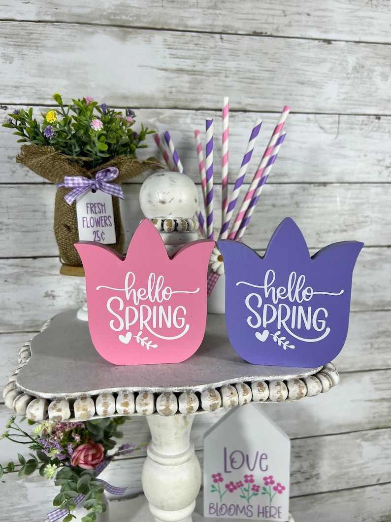 K&ouml;nnte beinhalten: Zwei h&ouml;lzerne tulpenf&ouml;rmige Schilder mit dem Text "hello spring" in wei&szlig;en Buchstaben. Die Schilder sind in rosa und lila bemalt und haben ein Herz- und Blattdesign. Die Schilder stehen auf einem wei&szlig;en, mehrstufigen St&auml;nder mit einem Schild, auf dem "love blooms here" steht.