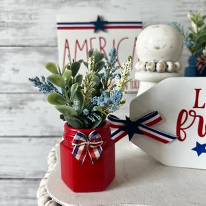 Pode incluir: Um pequeno frasco vermelho com um la&ccedil;o vermelho, branco e azul est&aacute; cheio de um ramo de vegeta&ccedil;&atilde;o artificial azul e verde. O frasco est&aacute; sentado em uma bandeja branca de v&aacute;rios n&iacute;veis com uma borda de contas de madeira brancas.