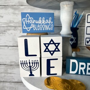 May include: White wooden blocks spelling out "LOVE" with a blue Star of David, menorah, and dreidel. The blocks are on a white tiered tray with gold chocolate coins and a sign that says "Hanukkah Blessings".