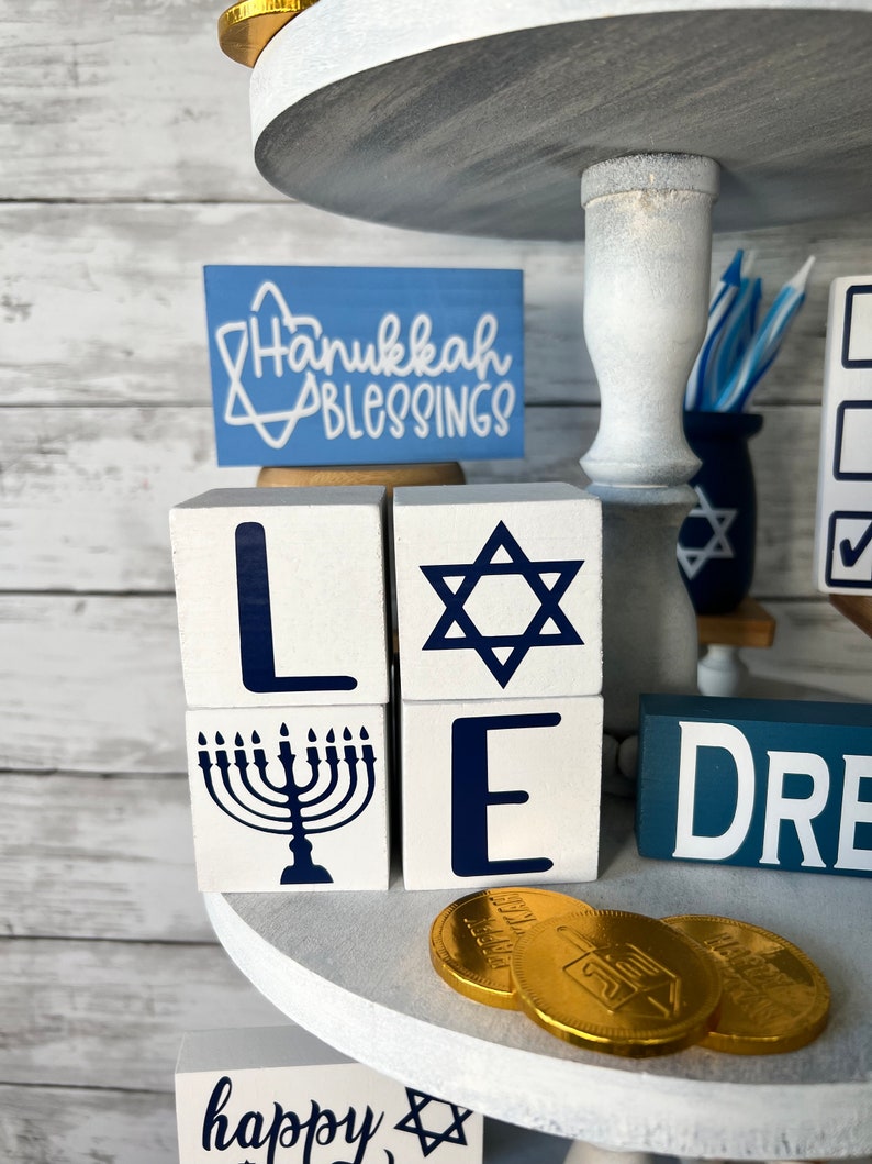 May include: White wooden blocks spelling out "LOVE" with a blue Star of David, menorah, and dreidel. The blocks are on a white tiered tray with gold chocolate coins and a sign that says "Hanukkah Blessings".