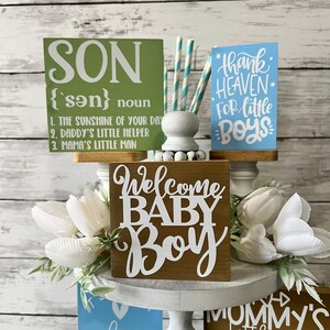 K&ouml;nnte beinhalten: Ein mehrst&ouml;ckiger Kuchenst&auml;nder mit verschiedenen Holzschilden mit Baby-Jungen-Motiven. Die Schilder sind mit wei&szlig;en Buchstaben verziert und beinhalten die S&auml;tze "Son", "Welcome Baby Boy", "Thank Heaven for Little Boys", "Mommy's Little Man Daddy's Best Bud" und "It's a Boy".