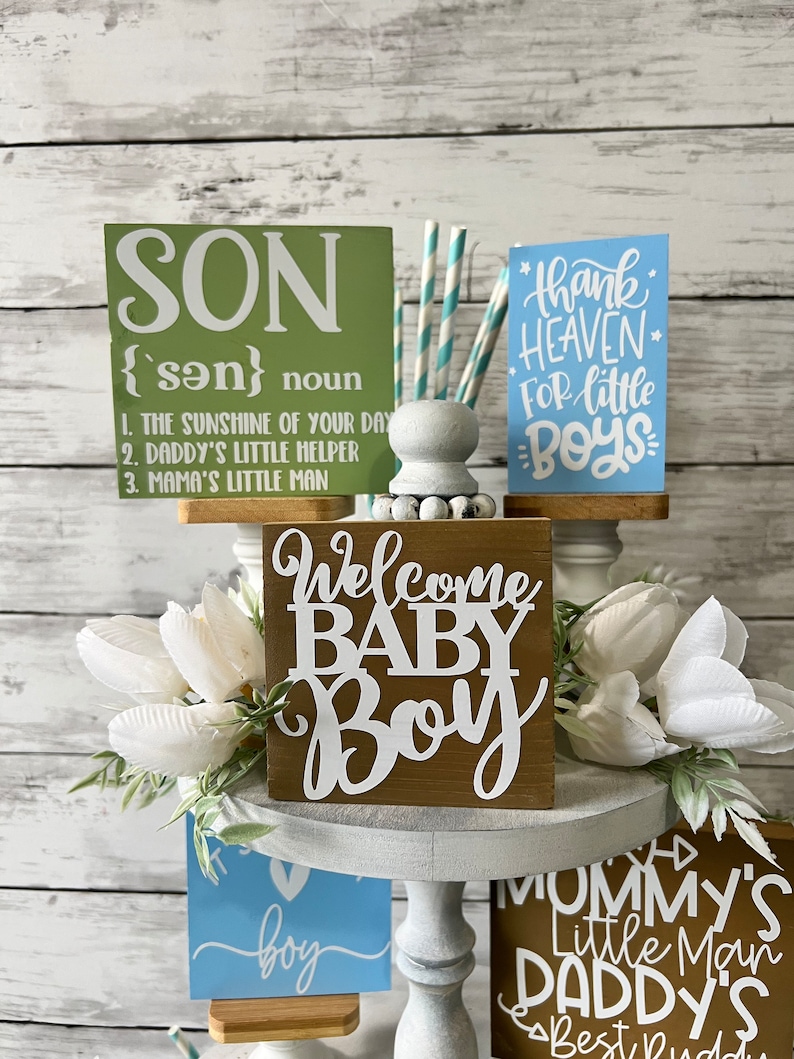 K&ouml;nnte beinhalten: Ein mehrst&ouml;ckiger Kuchenst&auml;nder mit verschiedenen Holzschilden mit Baby-Jungen-Motiven. Die Schilder sind mit wei&szlig;en Buchstaben verziert und beinhalten die S&auml;tze "Son", "Welcome Baby Boy", "Thank Heaven for Little Boys", "Mommy's Little Man Daddy's Best Bud" und "It's a Boy".