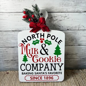 May include: White wooden chopping board with a red bow and green holly sprig. The board has the text "North Pole Milk & Cookie Company Baking Santa's Favourite Since 1896" in black and red lettering.
