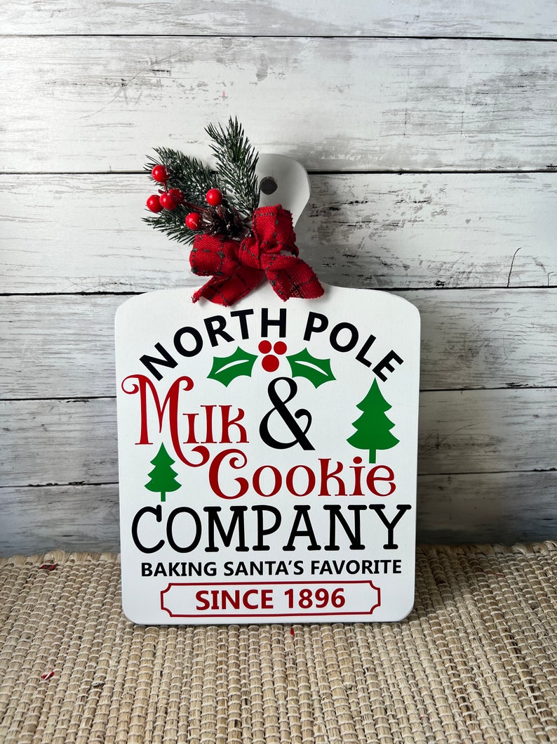 May include: White wooden chopping board with a red bow and green holly sprig. The board has the text "North Pole Milk & Cookie Company Baking Santa's Favourite Since 1896" in black and red lettering.