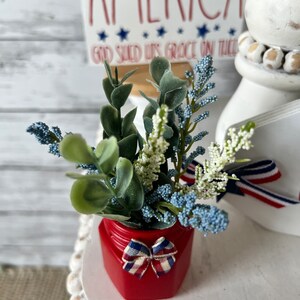 Pode incluir: Um frasco vermelho com um la&ccedil;o vermelho, branco e azul cont&eacute;m um buqu&ecirc; de flores azuis e brancas e folhas verdes. O frasco est&aacute; sentado em um suporte branco de v&aacute;rios n&iacute;veis com contas brancas. Uma placa com o texto "America God Shed His Grace On Thee" est&aacute; no fundo.