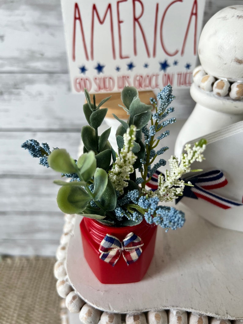 Pode incluir: Um frasco vermelho com um la&ccedil;o vermelho, branco e azul cont&eacute;m um buqu&ecirc; de flores azuis e brancas e folhas verdes. O frasco est&aacute; sentado em um suporte branco de v&aacute;rios n&iacute;veis com contas brancas. Uma placa com o texto "America God Shed His Grace On Thee" est&aacute; no fundo.
